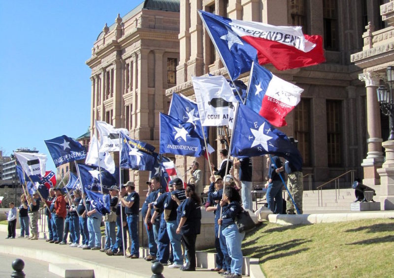 Supporters of the Texas Nationalist Movement hold flags on the steps of the Texas Capitol. The flags say "Independence," "Texas," and "Come and Take It," with a picture of a cannon. One Confederate flag is seen in the background.